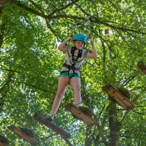 Tree Top Walk at Castlecomer Discovery Park A person wearing a helmet and harness crosses a wooden rope bridge in the Skywalk Challenge adventure park, surrounded by green trees. Another person waits on a platform to the left.