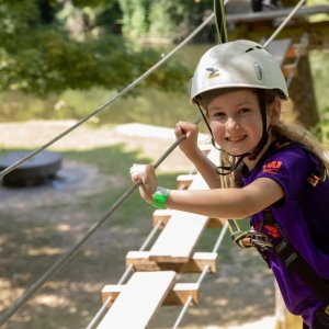 Tree Top Walk at Castlecomer Discovery Park A young girl wearing a helmet and harness smiles whilst crossing the Skywalk Challenge rope bridge in an outdoor adventure park, surrounded by trees and sunlight.