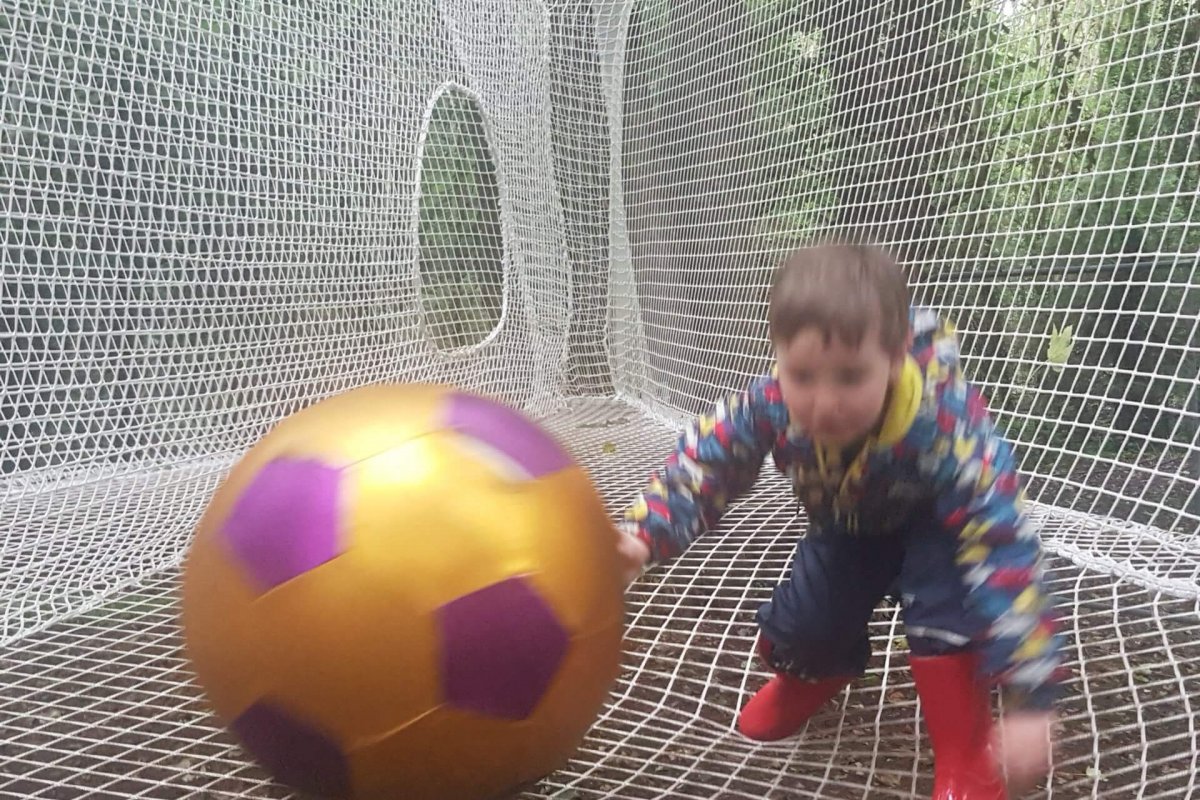 A young child in colourful clothes and red boots enjoys a Junior Woodland Adventure, playing on a white net structure outdoors and reaching for a large gold and purple football amid trees and greenery.