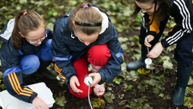 primary – invertebrate collection – 6.Still001 Three children in outdoor clothing crouch on the ground, using scientific equipment to examine soil or plants. The damp, leafy area suggests a hands-on nature or science activity, perfect for school trips looking to inspire curiosity outdoors.