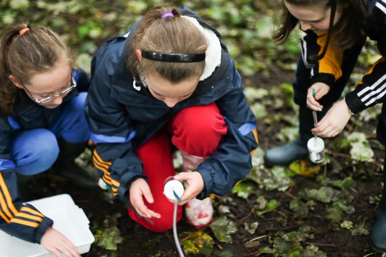 primary – invertebrate collection – 6.Still001 Three children in outdoor clothing crouch on the ground, using scientific equipment to examine soil or plants. The damp, leafy area suggests a hands-on nature or science activity, perfect for school trips looking to inspire curiosity outdoors.